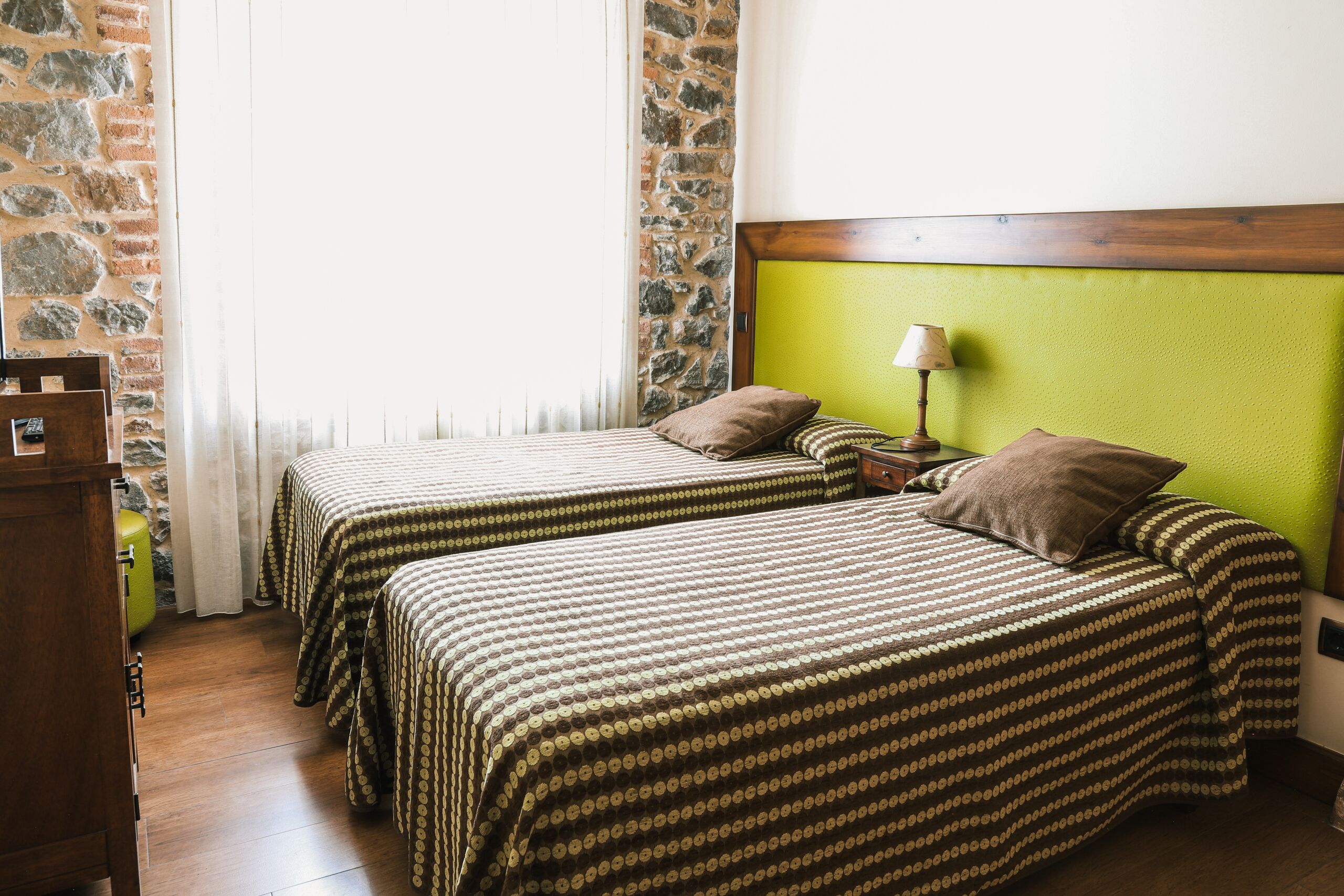 A room with two beds, a wooden headboard, a nightstand with a lamp, and a stone wall.