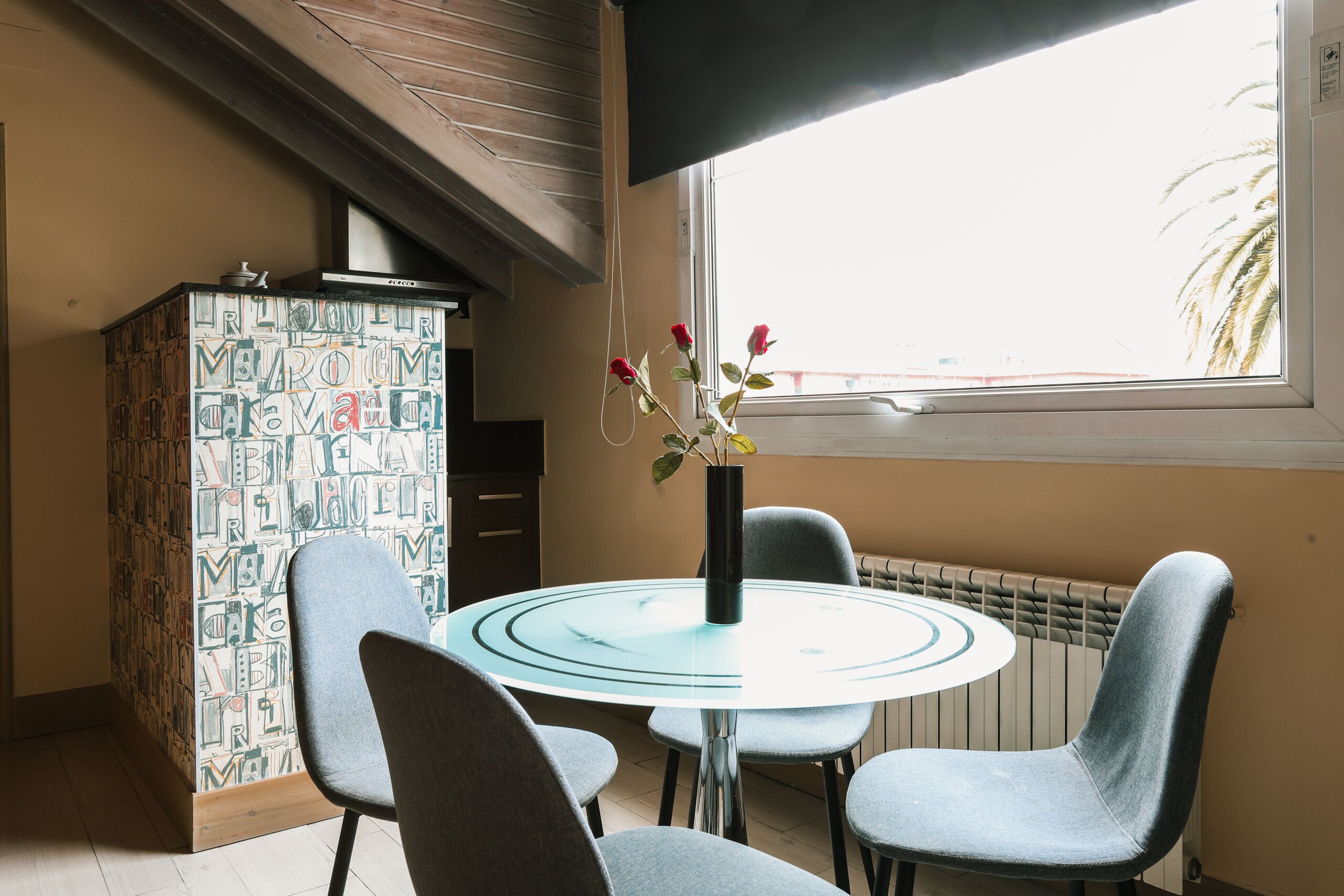 Un coin repas comprenant une table ronde en verre, quatre chaises grises, un vase avec des fleurs rouges et un buffet à motifs.