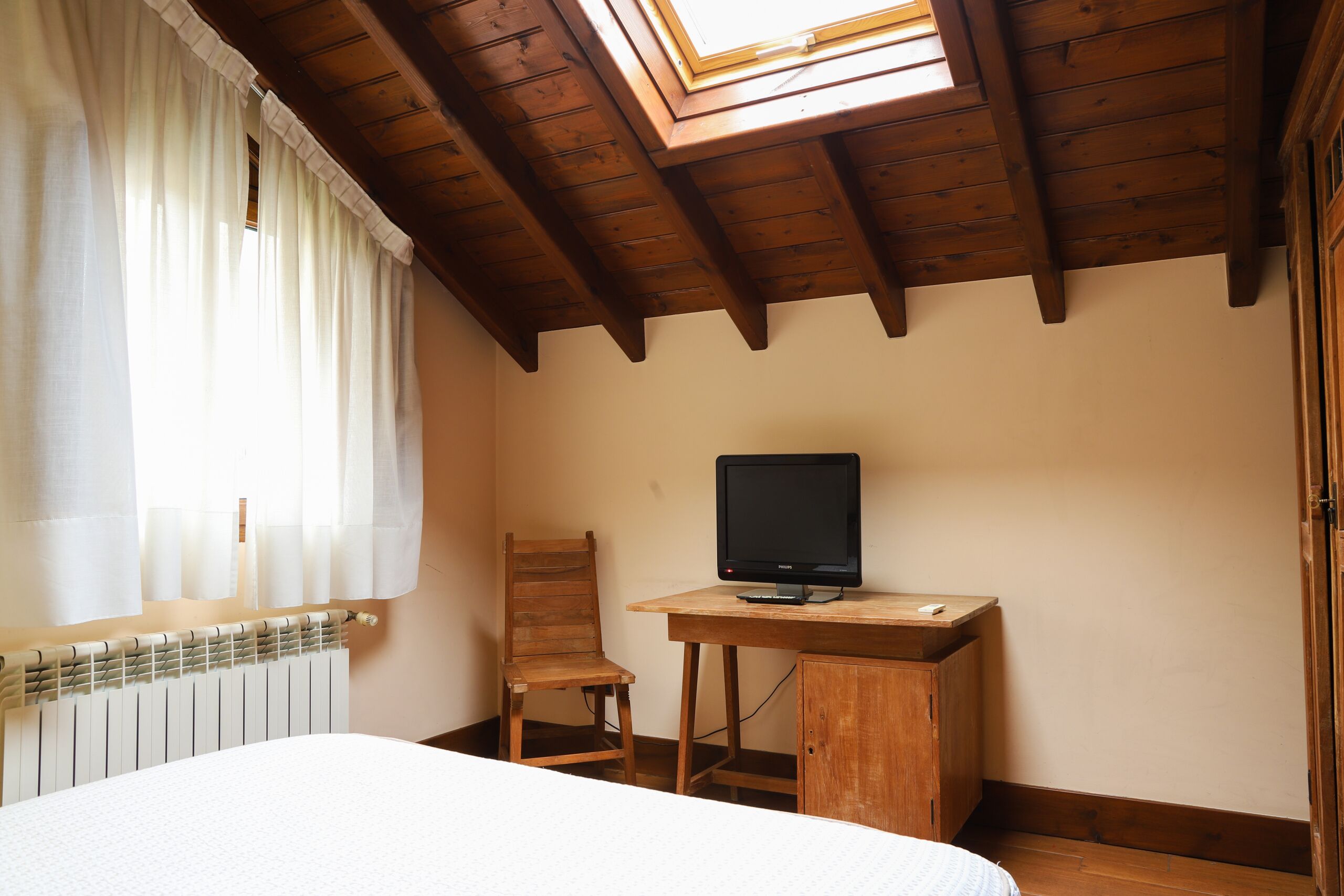 A bedroom with a wooden desk, a chair, a bed, a window with curtains, and a skylight.
