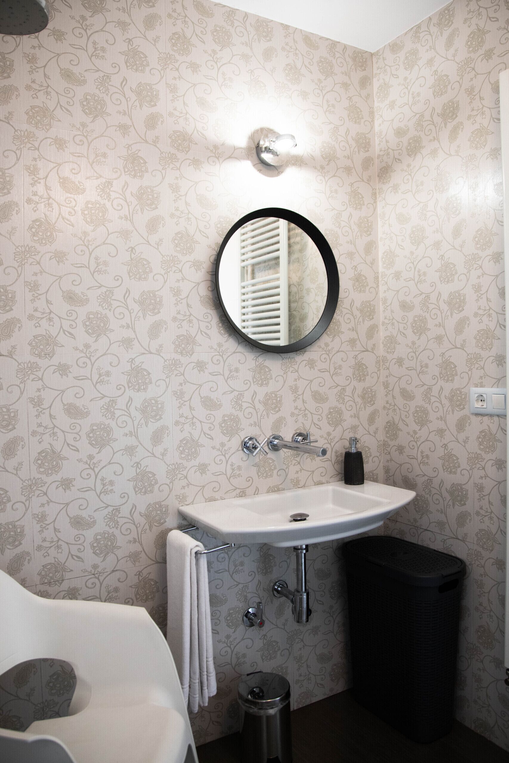A bathroom with a round mirror, white sink, and a black trash bin.