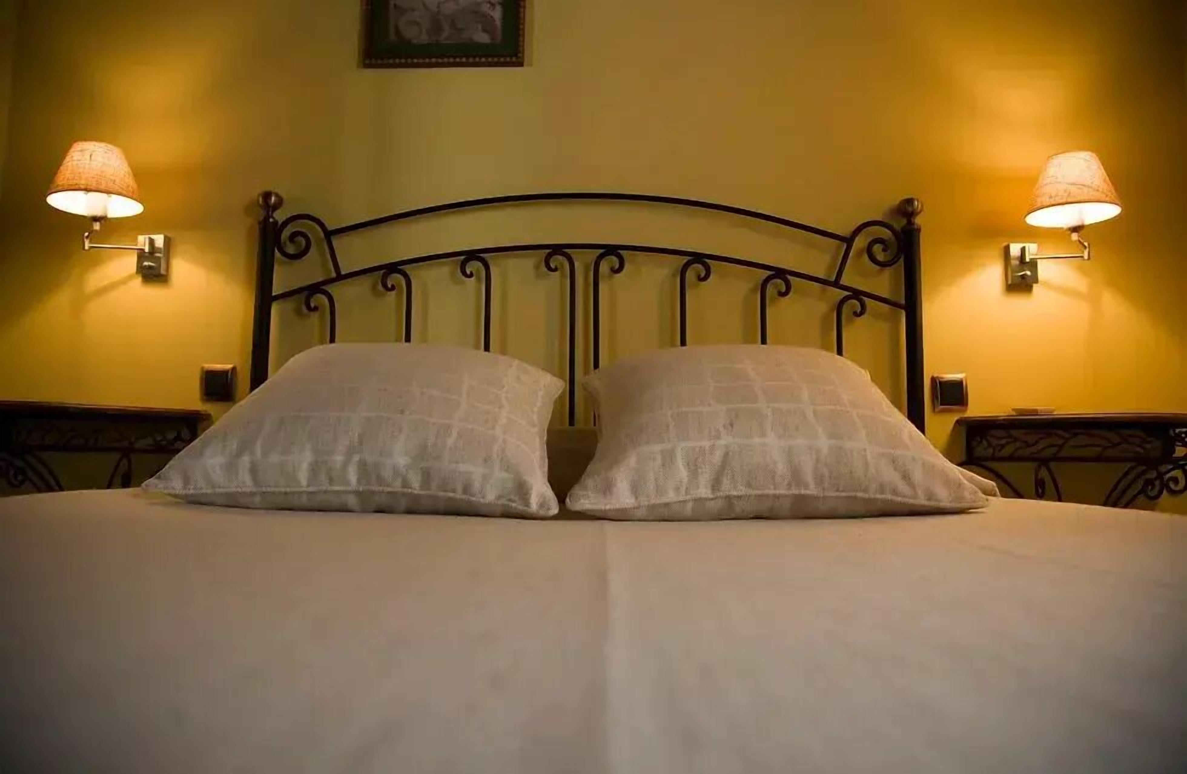 Una cama con dos almohadas y cabecera de metal. Hay dos lámparas de pared a cada lado de la cama.
