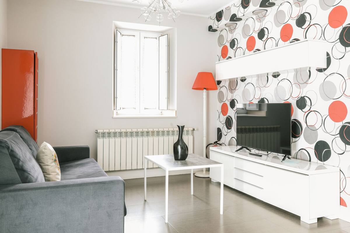 Un salon moderne avec un canapé gris, une table basse blanche, un téléviseur posé sur un meuble blanc et un mur orné d’un motif.