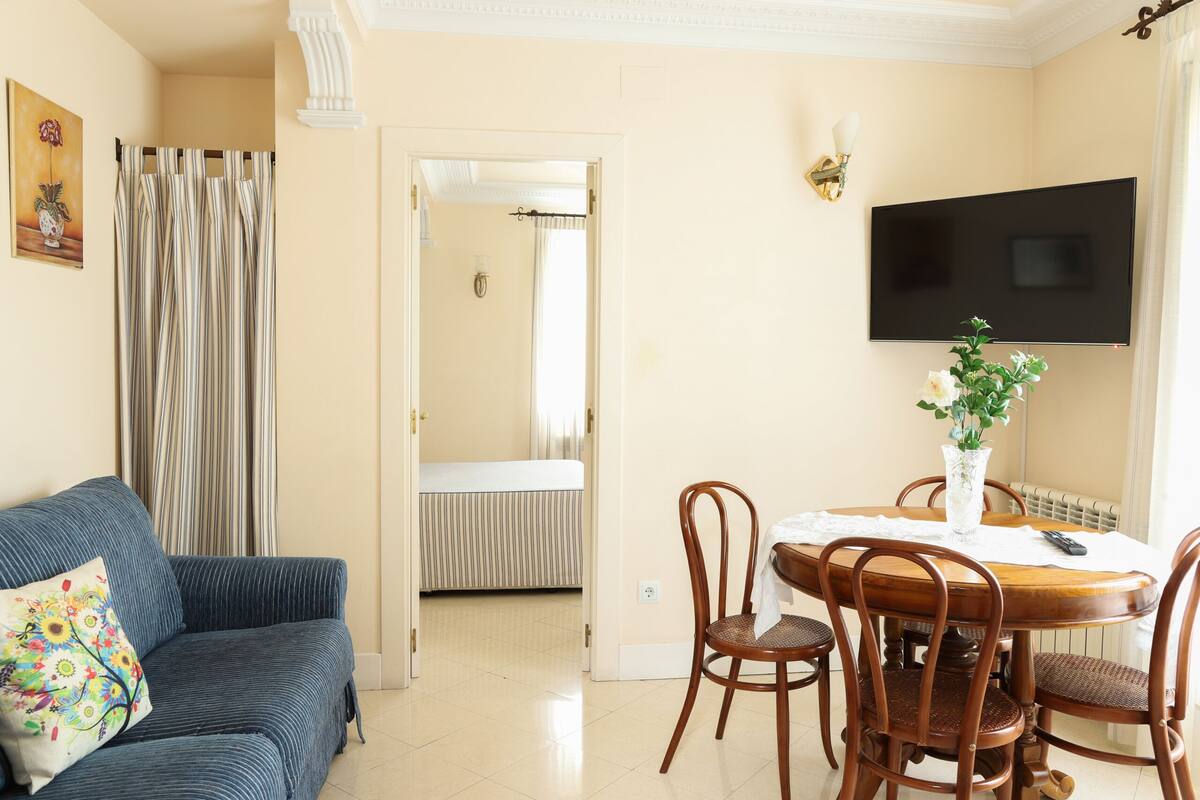 Un salon comprenant un canapé, une table à manger avec des chaises, un téléviseur fixé au mur et une porte donnant sur une autre pièce.