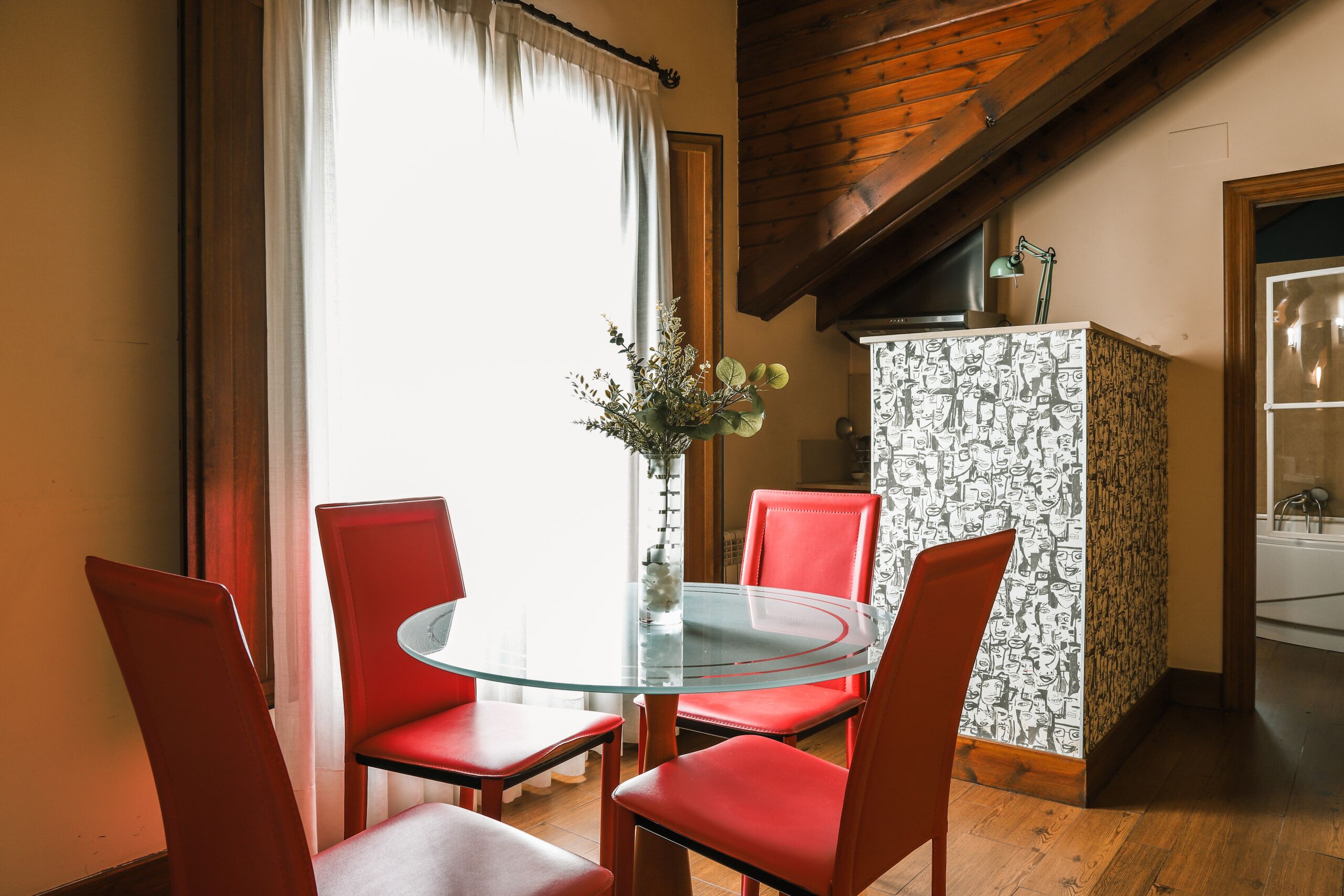Un coin repas avec une table en verre et des chaises rouges, un vase rempli de fleurs et un buffet à motifs.