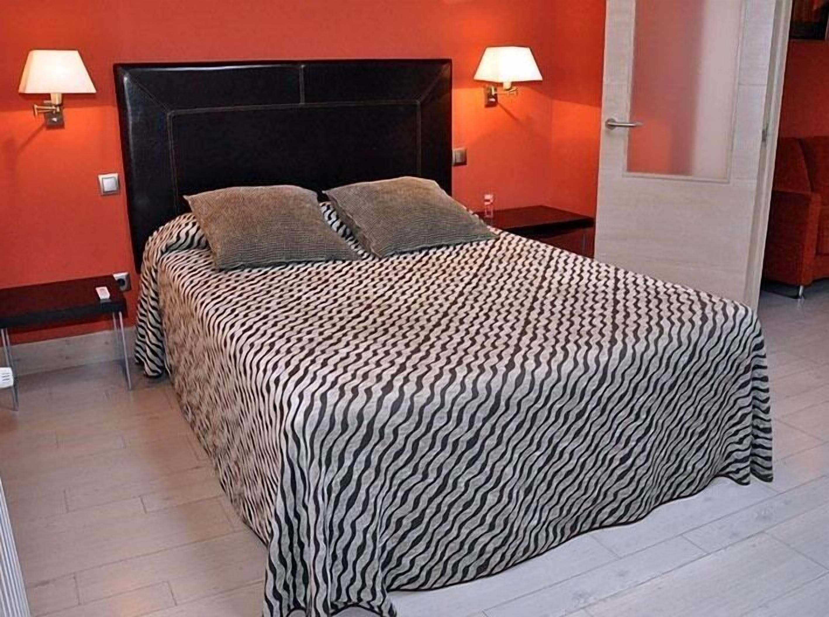 Una cama con colcha de estampado de cebra, dos almohadas y dos lámparas de noche en una habitación con paredes rojas y suelo de madera.