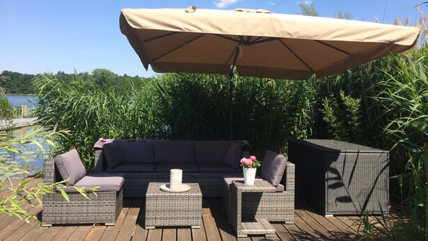 Ein Sitzbereich im Freien mit einer Couch, zwei Beistelltischen und einem kleinen Tisch im Freien, alles auf einer Holzterrasse mit Blick auf einen See und grüne Landschaft im Hintergrund.