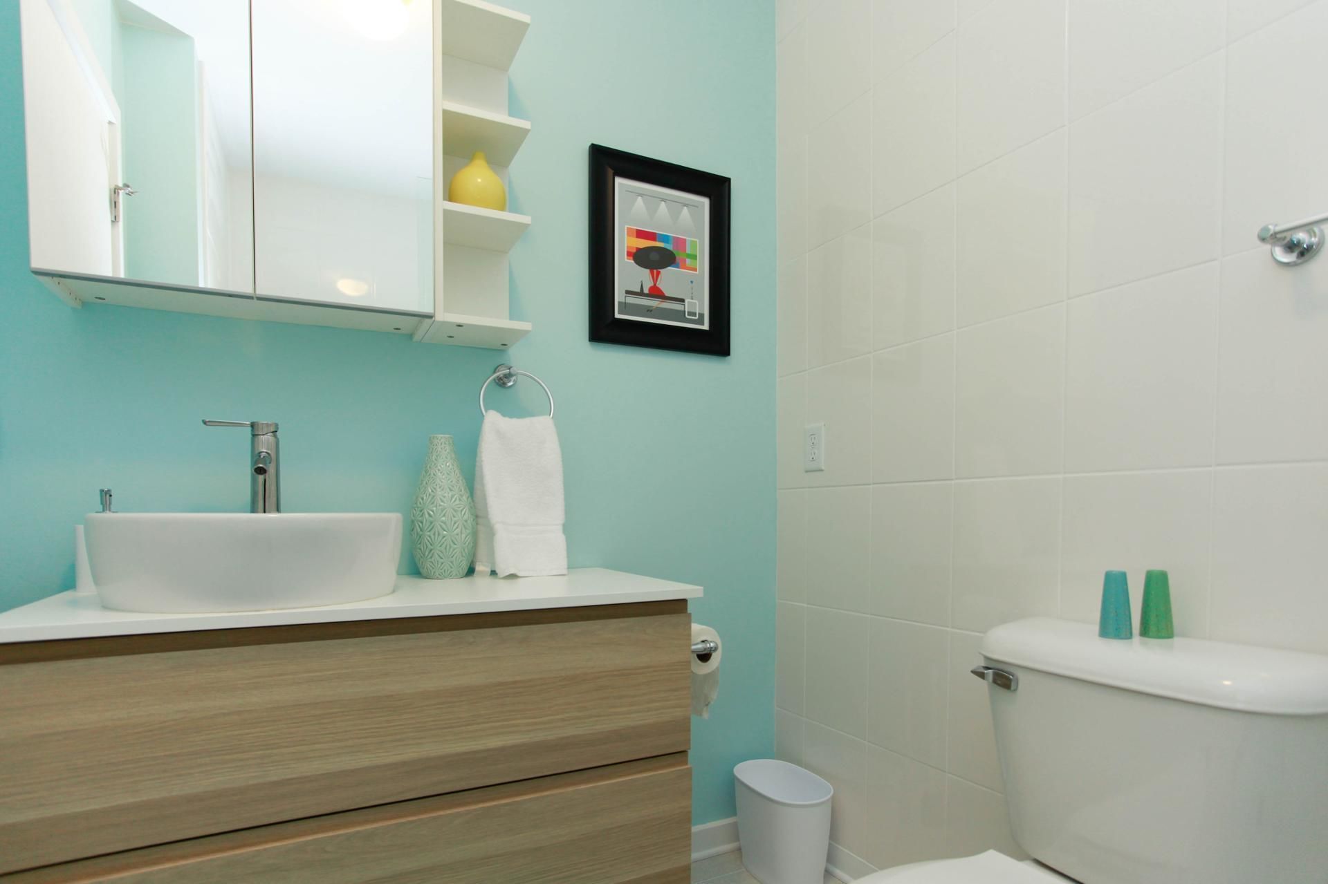 Ein Badezimmer mit weißem Waschbecken, Holz-Waschtisch, einem gerahmten Bild und einer Toilette mit zwei Flaschen auf dem Tank.