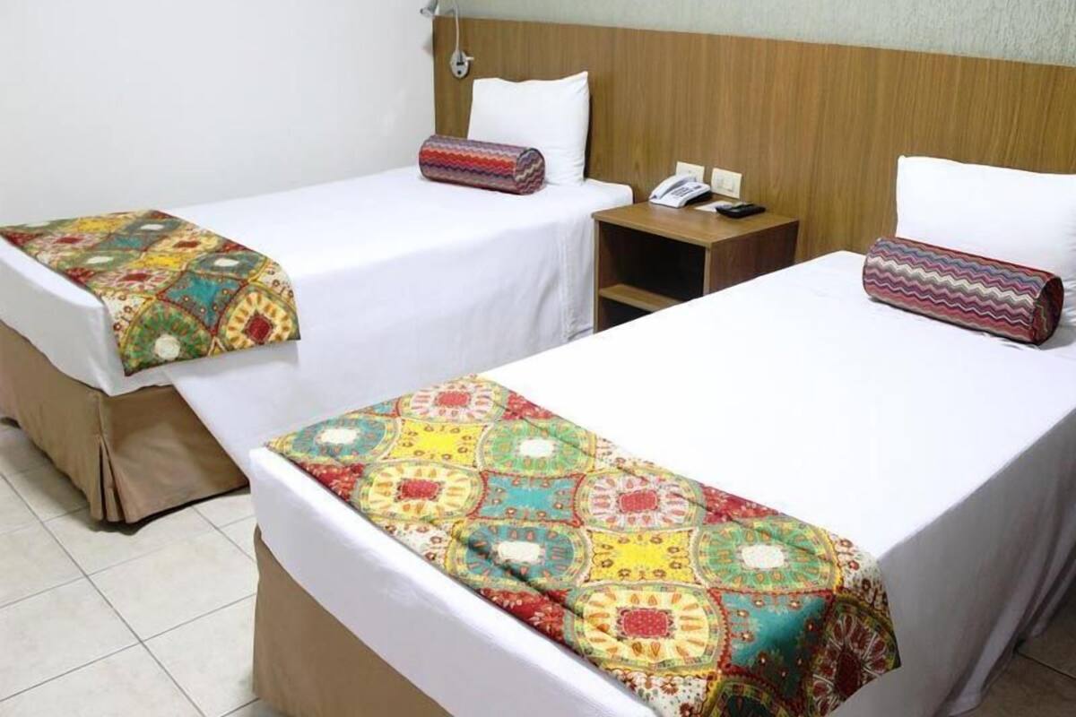 A hotel room with two single beds, each with a patterned bedspread and pillow, a bedside table with a phone and a lamp, and a wooden headboard.