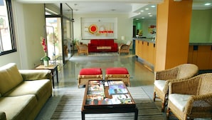 A hotel lobby with a red sofa, wicker chairs, a coffee table with magazines, and a reception desk. - Hotel Riviera (São José do Rio Preto)