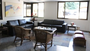 A modern living room with a black leather sofa, wicker chairs, a coffee table with magazines, and large windows offering a view of the street. - Hotel Riviera (São José do Rio Preto)