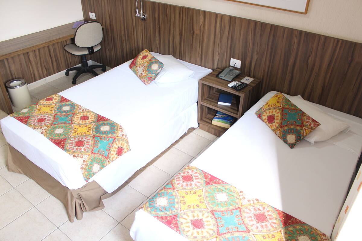 Two single beds with colorful bedding, a wooden headboard, a desk with a chair, and a bedside table with a phone and books.