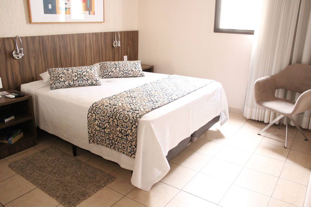 A neatly made bed with patterned bedding, a bedside table with a lamp, and a chair in a well-lit room with a window and curtains.