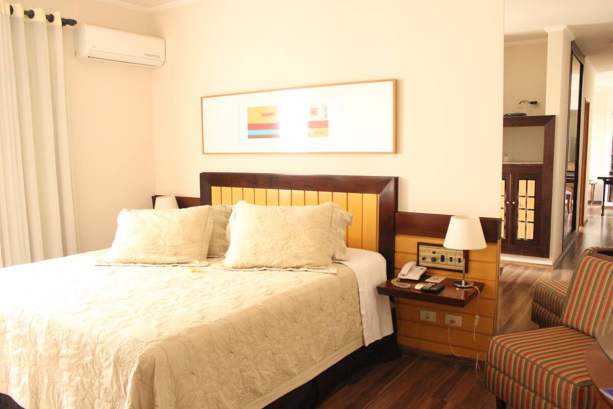 A hotel room with a large bed, bedside table with a lamp, a chair, and a framed picture on the wall.
