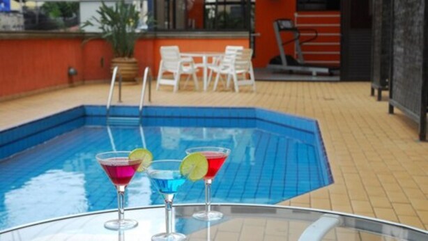 A swimming pool with three colorful drinks, a sign for 'RIVIERA', and outdoor seating. - Hotel Riviera (São José do Rio Preto)