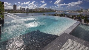 Una piscina con caída de agua, rodeada de sillones de descanso y sombrillas, con un horizonte urbano al fondo.