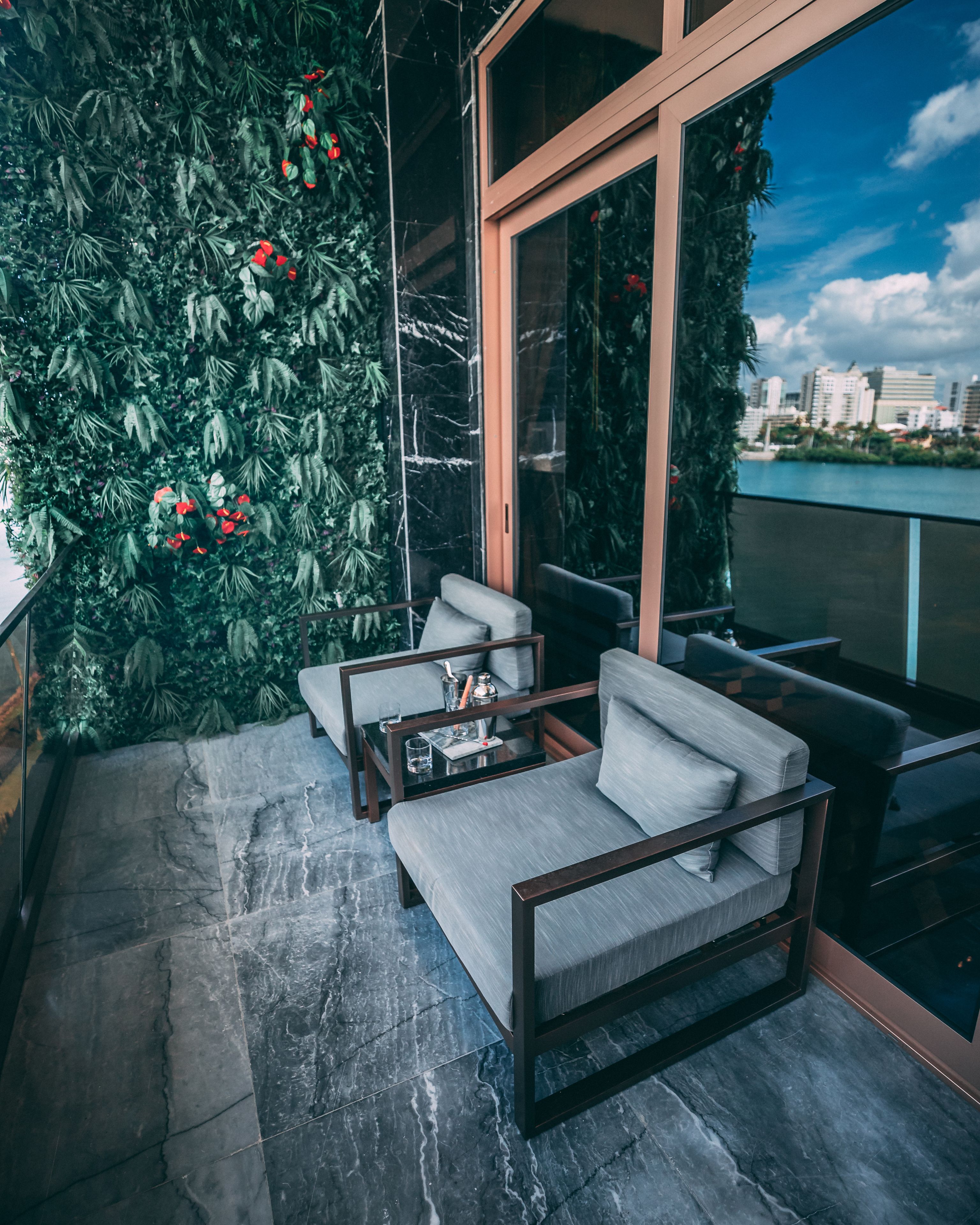 A modern balcony with a grey sofa, a small table, and a decorative wall with green foliage and red flowers.