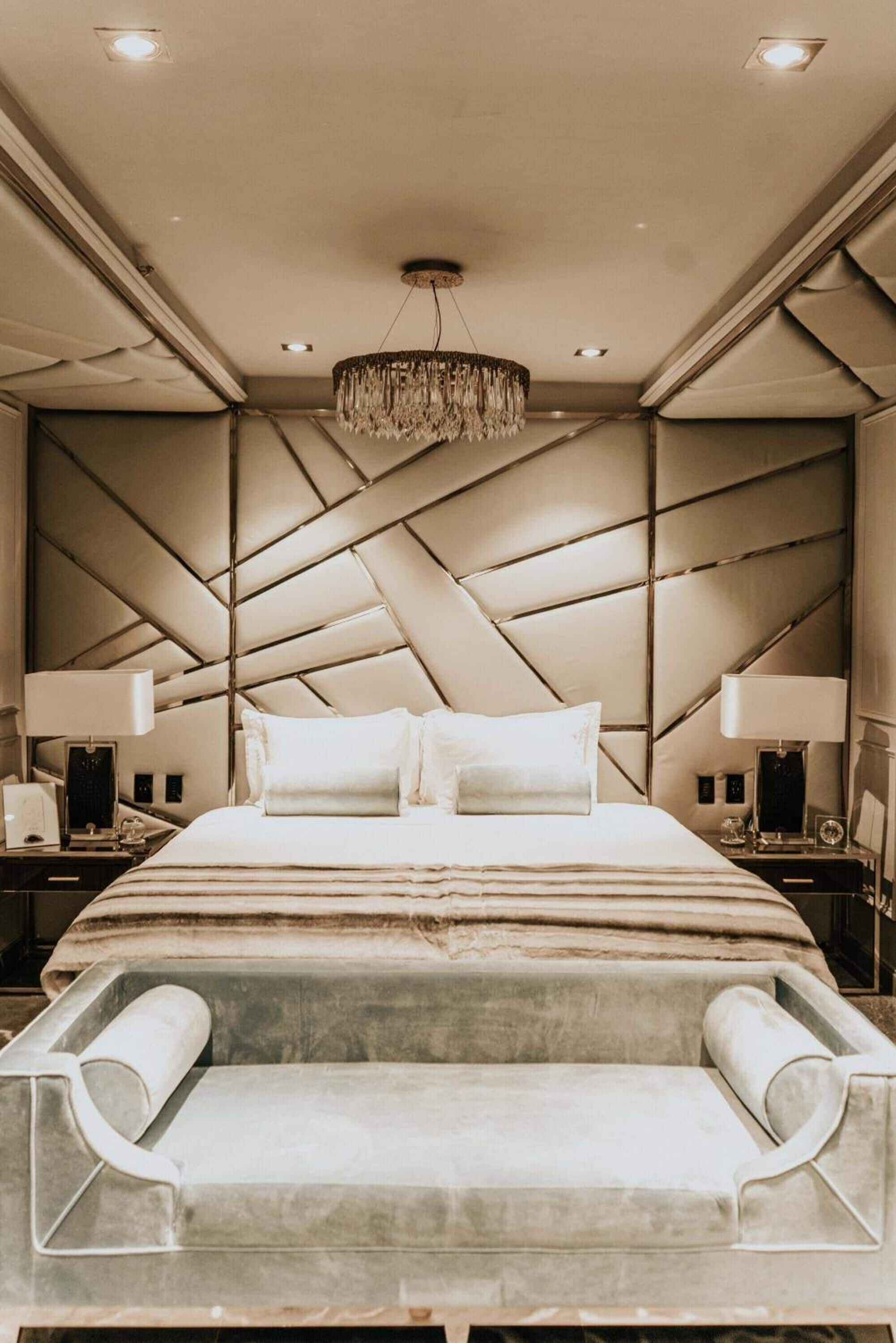 A modern hotel room with a large bed, a chandelier, and a unique geometric headboard.