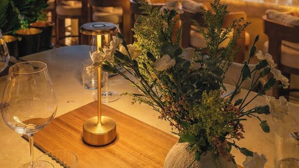Un restaurante con vistas al horizonte de la ciudad al anochecer, con una zona de bar con estantes llenos de botellas y una mesa con un jarrón de flores y copas de vino.