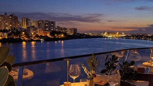 Una terraza con una mesa lista para cenar, con vistas a un paisaje urbano frente al mar al atardecer.