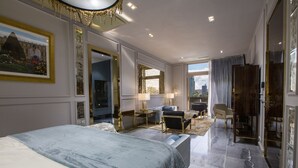 Amplia habitación de hotel con una cama grande, una zona de estar con sofá y sillones, un escritorio y vistas a la ciudad a través de amplias ventanas.