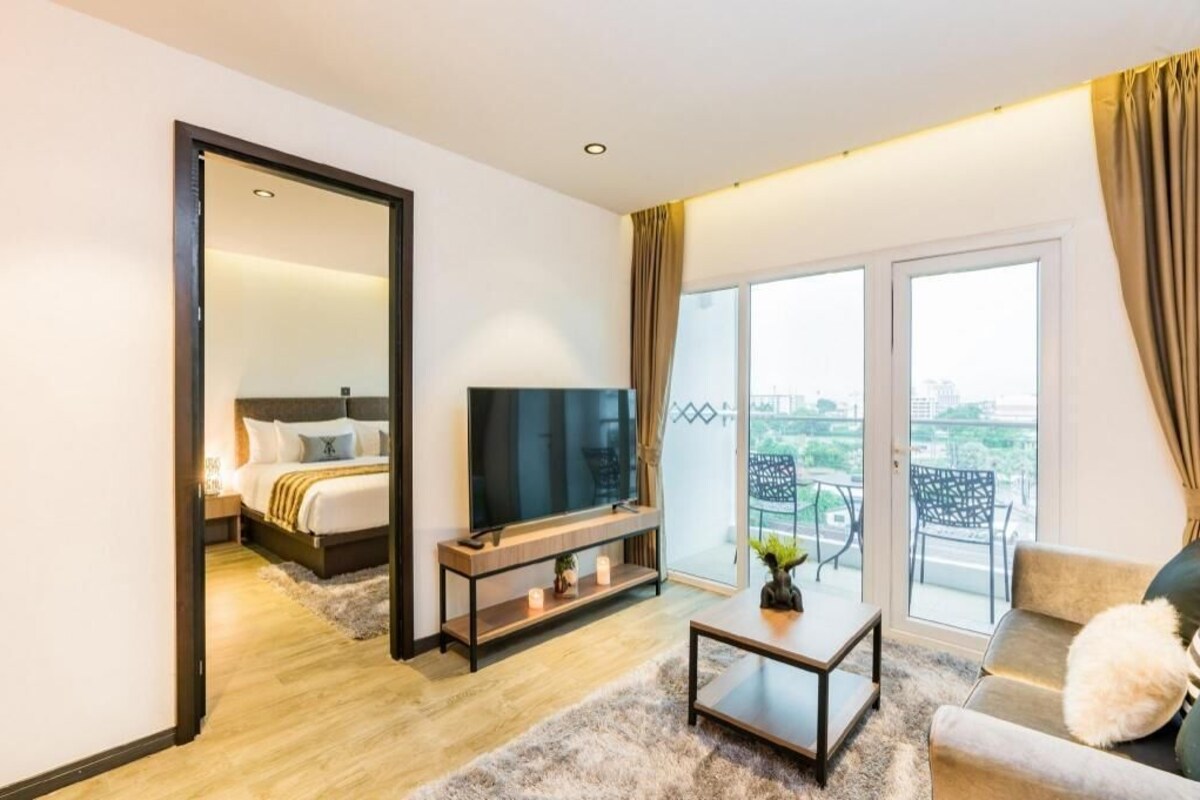 A modern hotel room with a bed, a TV, a sofa, a coffee table, and a balcony with outdoor furniture.