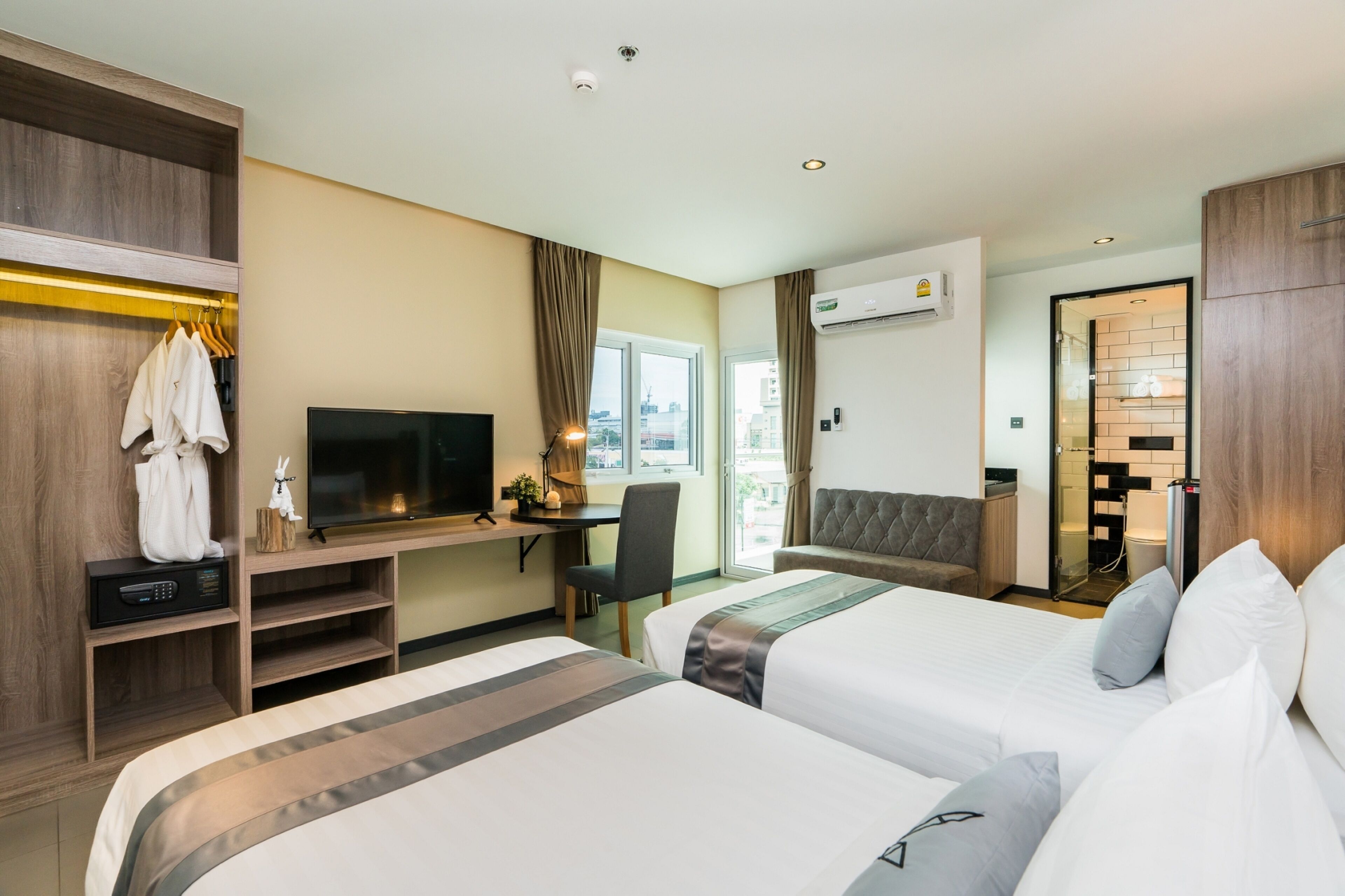 Habitación de hotel con dos camas, un escritorio con televisor, un armario y un baño visible a través de una puerta abierta.
