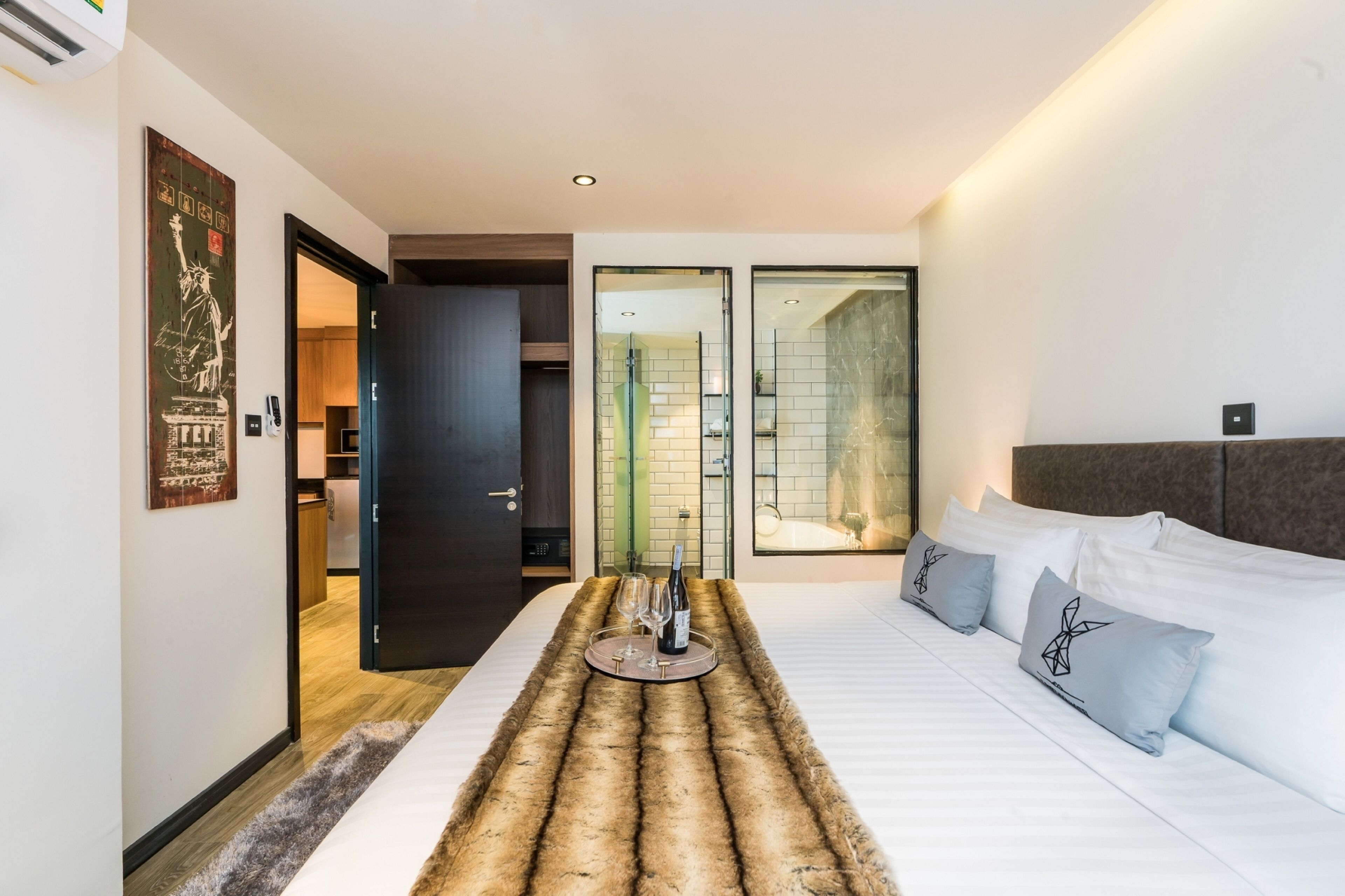 Ett modernt hotellrum med en säng, en fäll av fuskpäls, en minibar och ett badrum som syns genom en glasdörr.