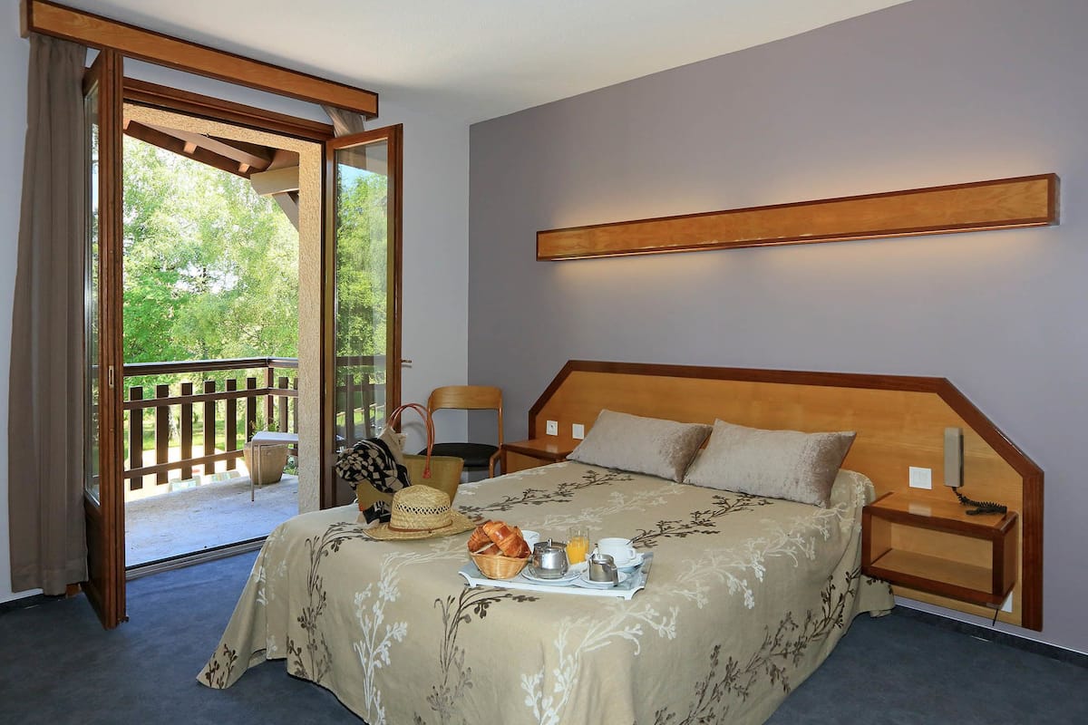Une chambre à coucher comprenant un lit, des tables de chevet, une commode, une fenêtre donnant sur un espace verdoyant et un balcon.
