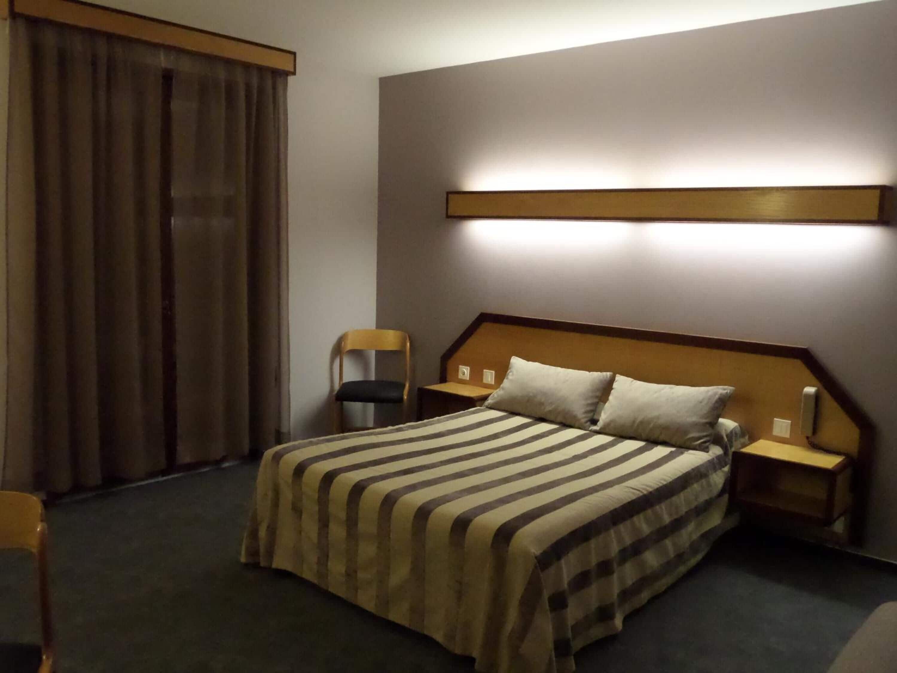 A hotel room with a bed, a chair, a nightstand, and a wall-mounted light.
