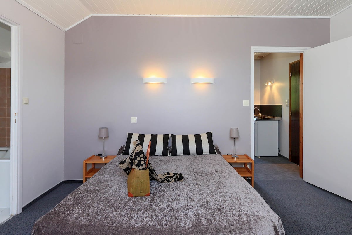 Une chambre à coucher comprenant un lit, deux tables de chevet, une commode, une lampe, un luminaire fixé au mur et une porte donnant sur une petite cuisine.