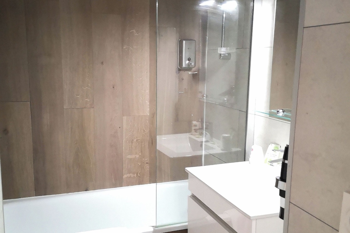 A modern bathroom with a wooden wall, a glass shower enclosure, a white sink, and a mirror.