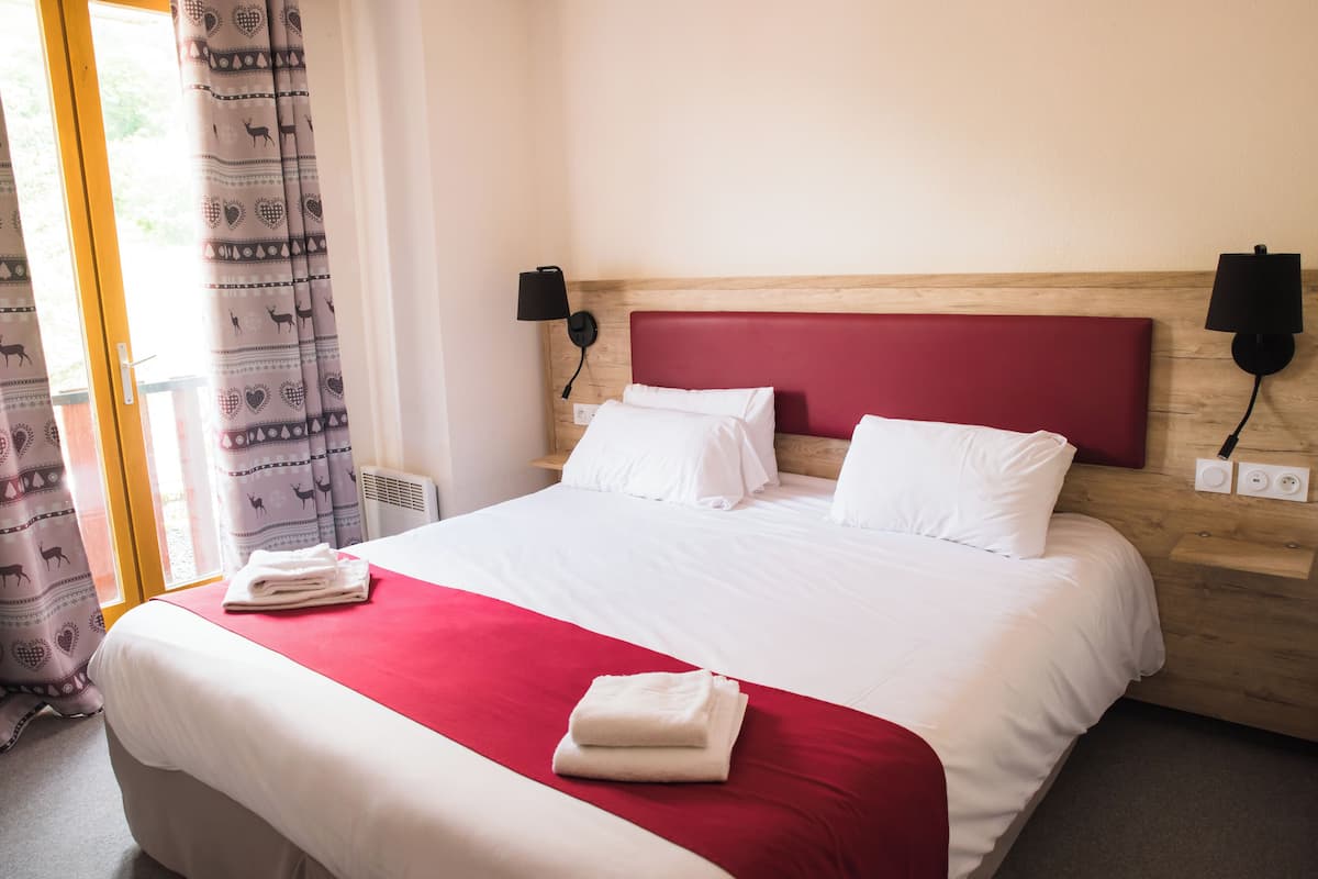 A neatly made bed with white and red bedding, two bedside lamps, and a window with patterned curtains.