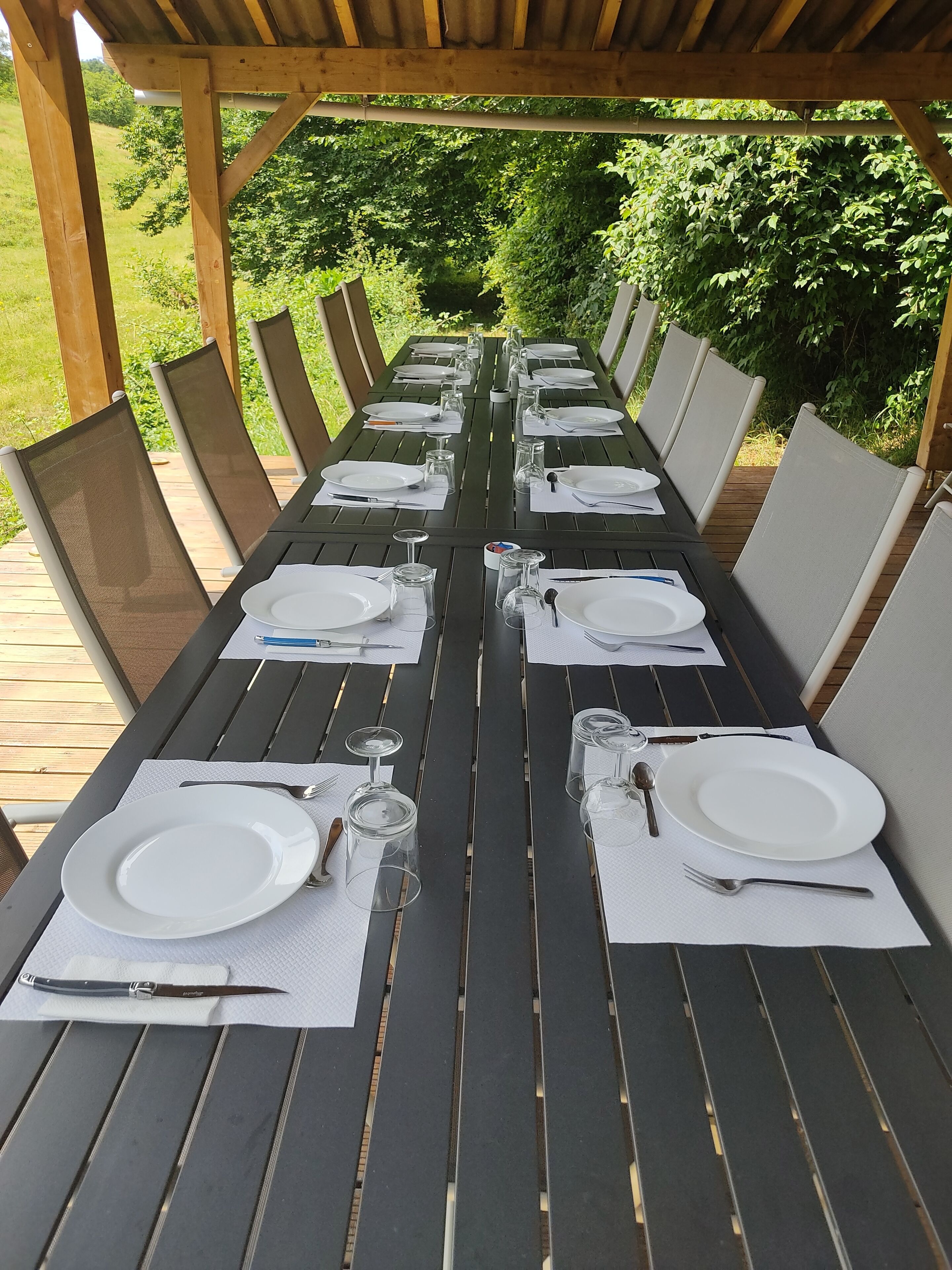 A long outdoor dining table set with white plates, cutlery, and glasses, surrounded by chairs with mesh backs.