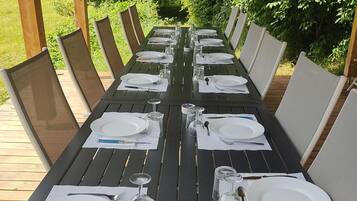 A long outdoor dining table set with white plates, cutlery, and glasses, surrounded by chairs with mesh backs.