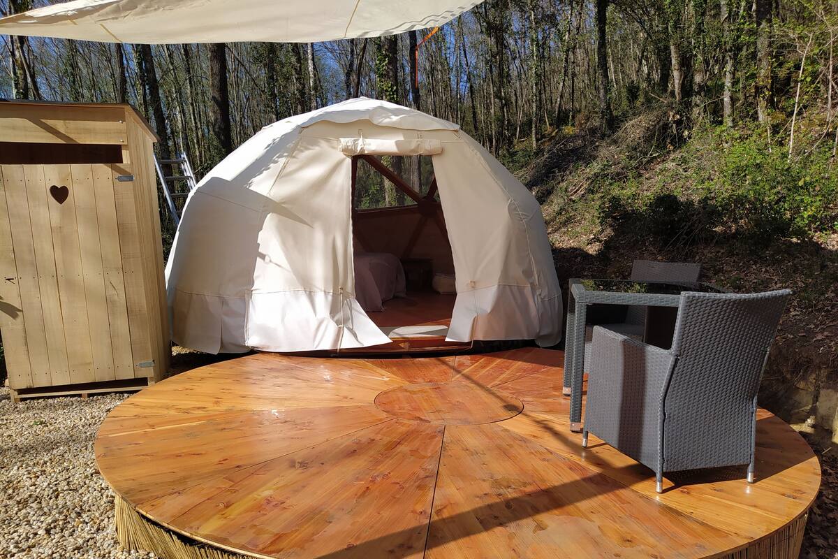 A round wooden table with a white tent and wicker chairs in a forest setting.