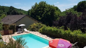 A rectangular swimming pool with a tiled deck, surrounded by lush greenery and a small building with a tiled roof. - La Maison de Tari (Vitrac)
