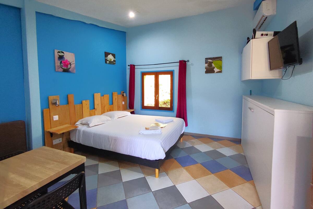 A bedroom with a bed, a desk, a chair, a window with curtains, a television, and a refrigerator.