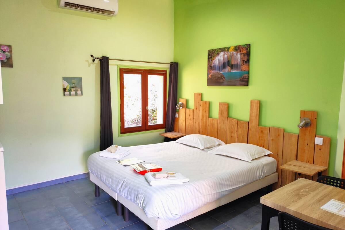 A room with a bed, a wooden headboard, a window with curtains, a painting on the wall, and a small table.