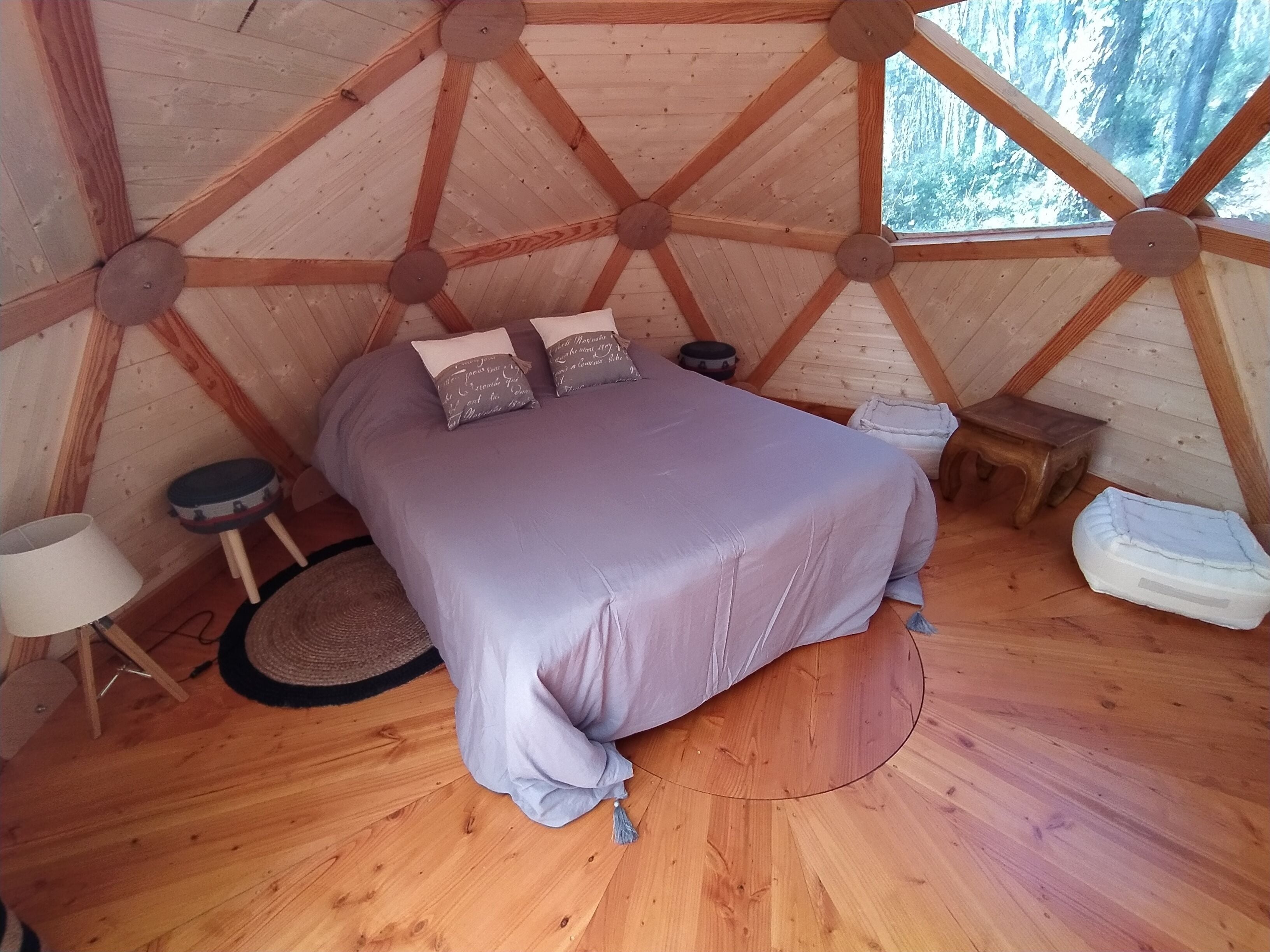 Un interior de madera cálido y circular, con una cama, almohadas, un taburete pequeño y una lámpara.