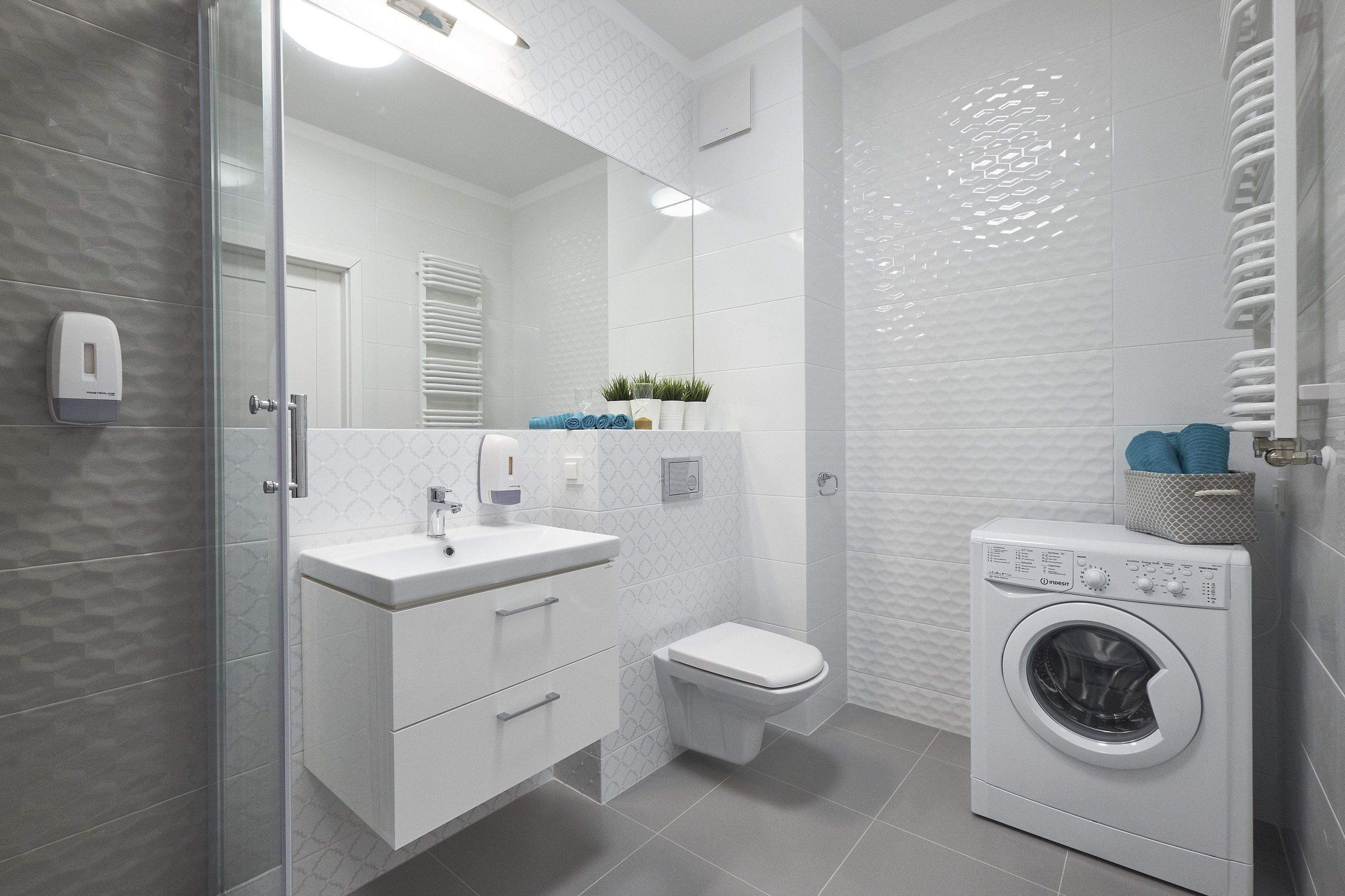 Et moderne badeværelse med brusekabine i glas, et hvidt håndvaskarrangement med spejl, toilet og en frontbetjent vaskemaskine.