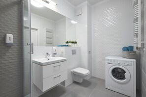 A modern bathroom with a glass shower enclosure, a white sink with a mirror, a toilet, and a front-loading washing machine. - Good Time Apartment (Warsaw)
