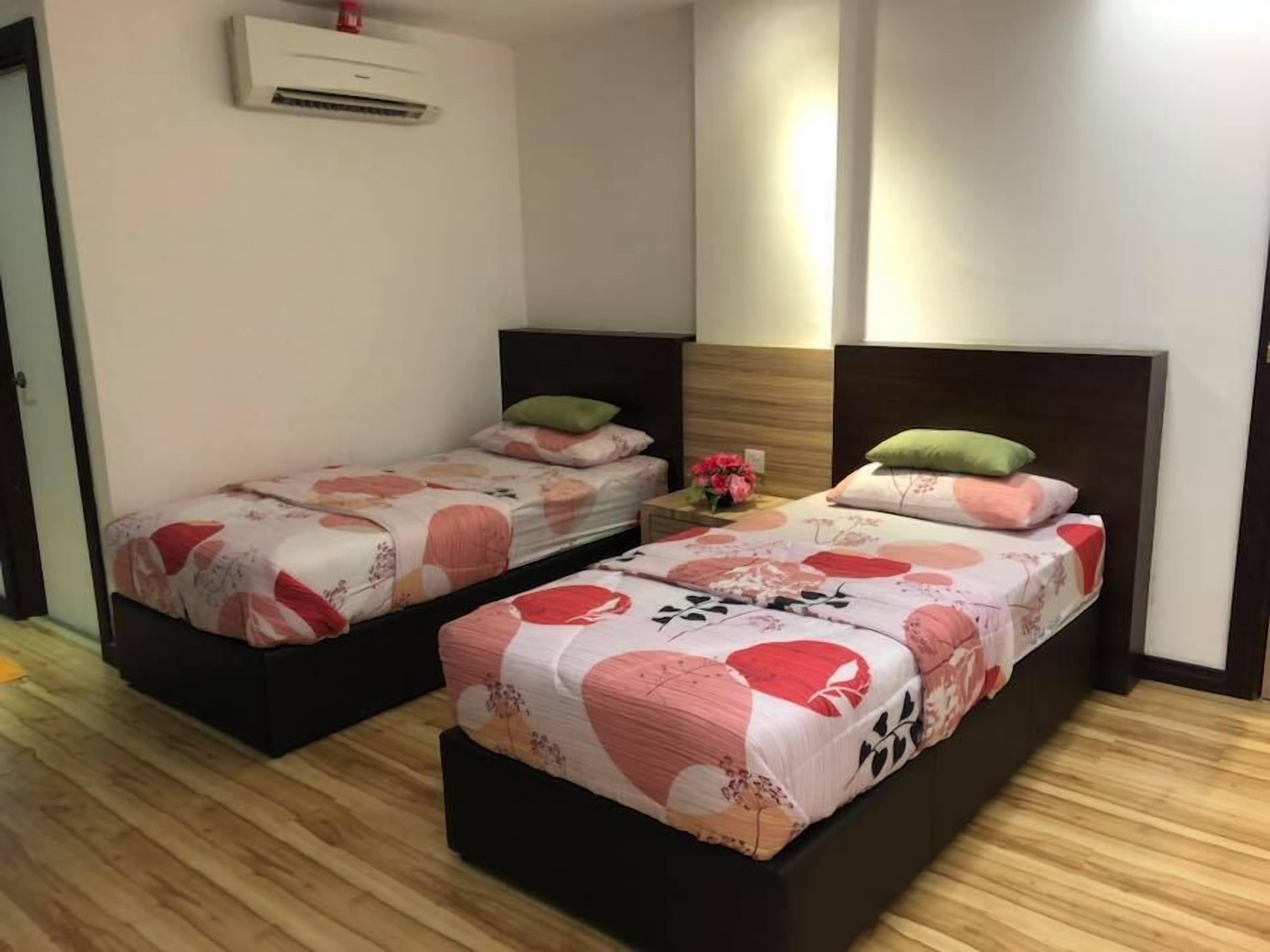 Habitación de hotel con dos camas individuales, piso de madera, unidad de aire acondicionado, mesita de noche con un florero y un espejo en la pared.