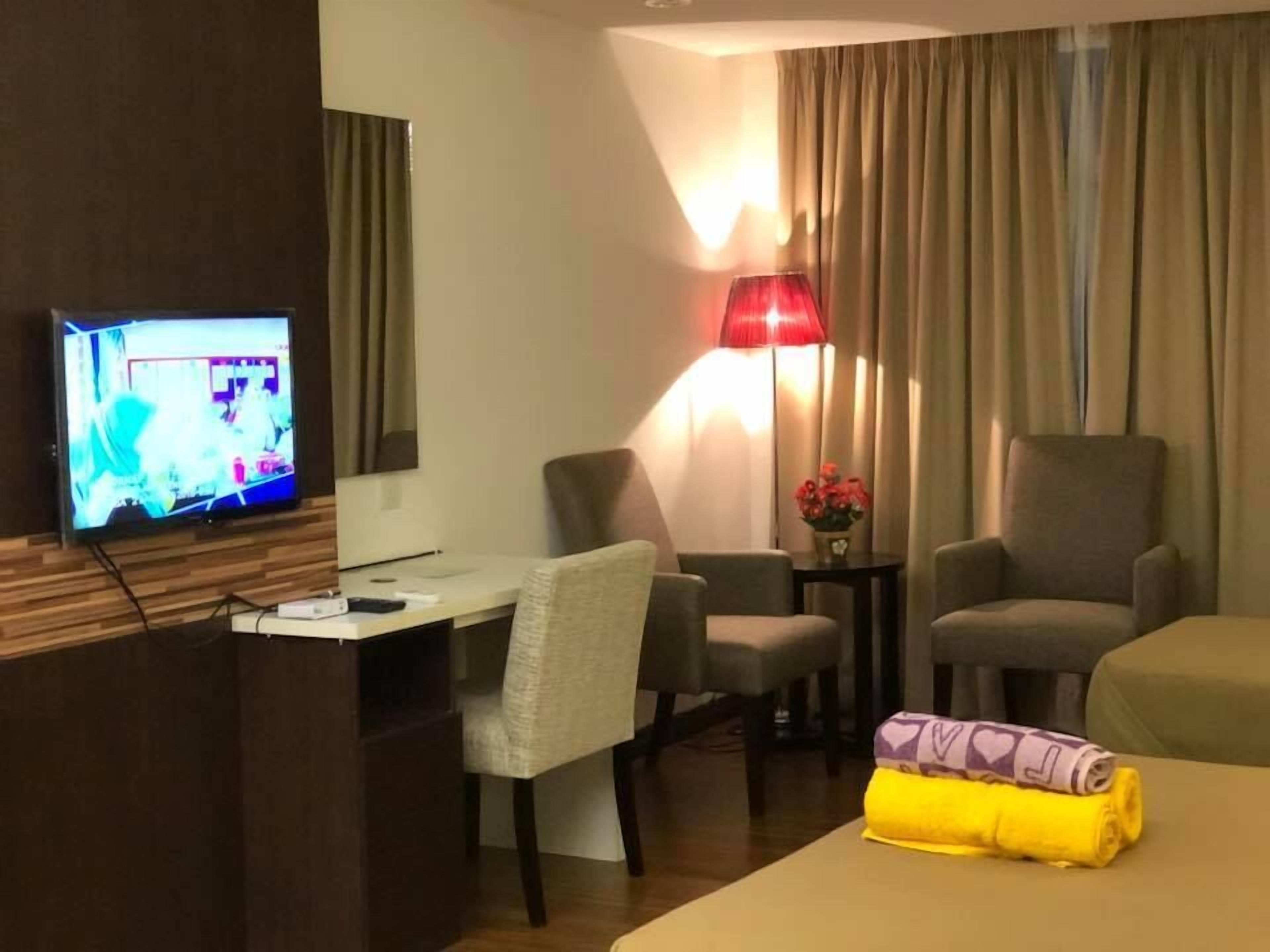 A hotel room with a TV, two armchairs, a desk, a lamp, and a bed with a rolled-up towel.