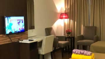 A hotel room with a TV, two armchairs, a desk, a lamp, and a bed with a rolled-up towel.