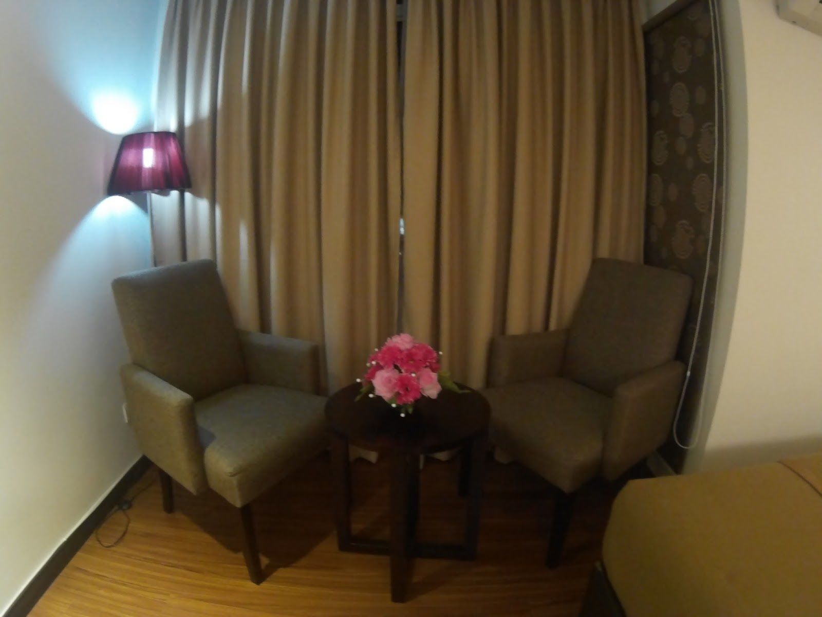 Une chambre d’hôtel avec deux fauteuils, une petite table sur laquelle est posé un vase de fleurs, une lampe et un rideau.