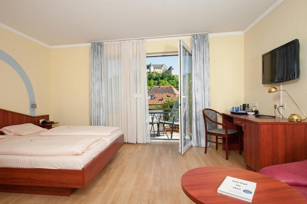 A hotel room with a bed, a desk, a chair, a TV, and a view of a building through a window.