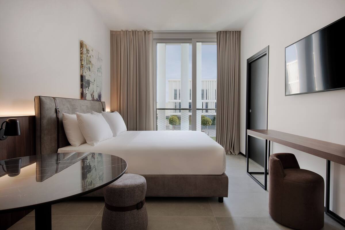A modern hotel room with a large bed, a flat-screen TV, a desk, and a view of the city through large windows.