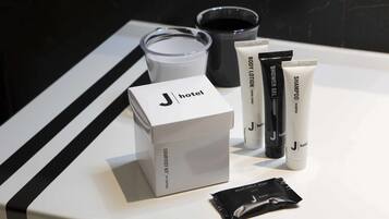 Un baño de hotel con encimera blanca, caja de artículos de cortesía, loción corporal, gel de ducha, champú y un paquete de productos de aseo.
