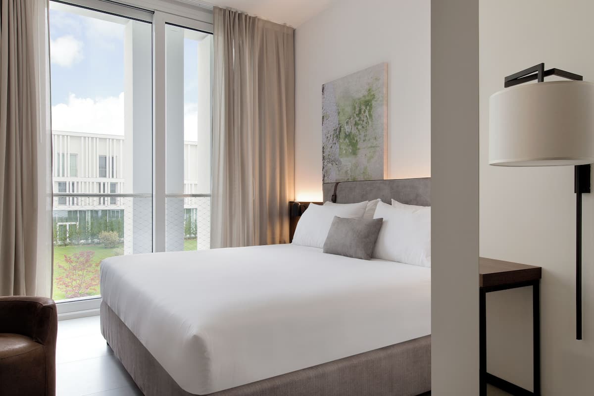 A hotel room with a large bed, a view of the outdoors, a painting on the wall, and a lamp on a bedside table.