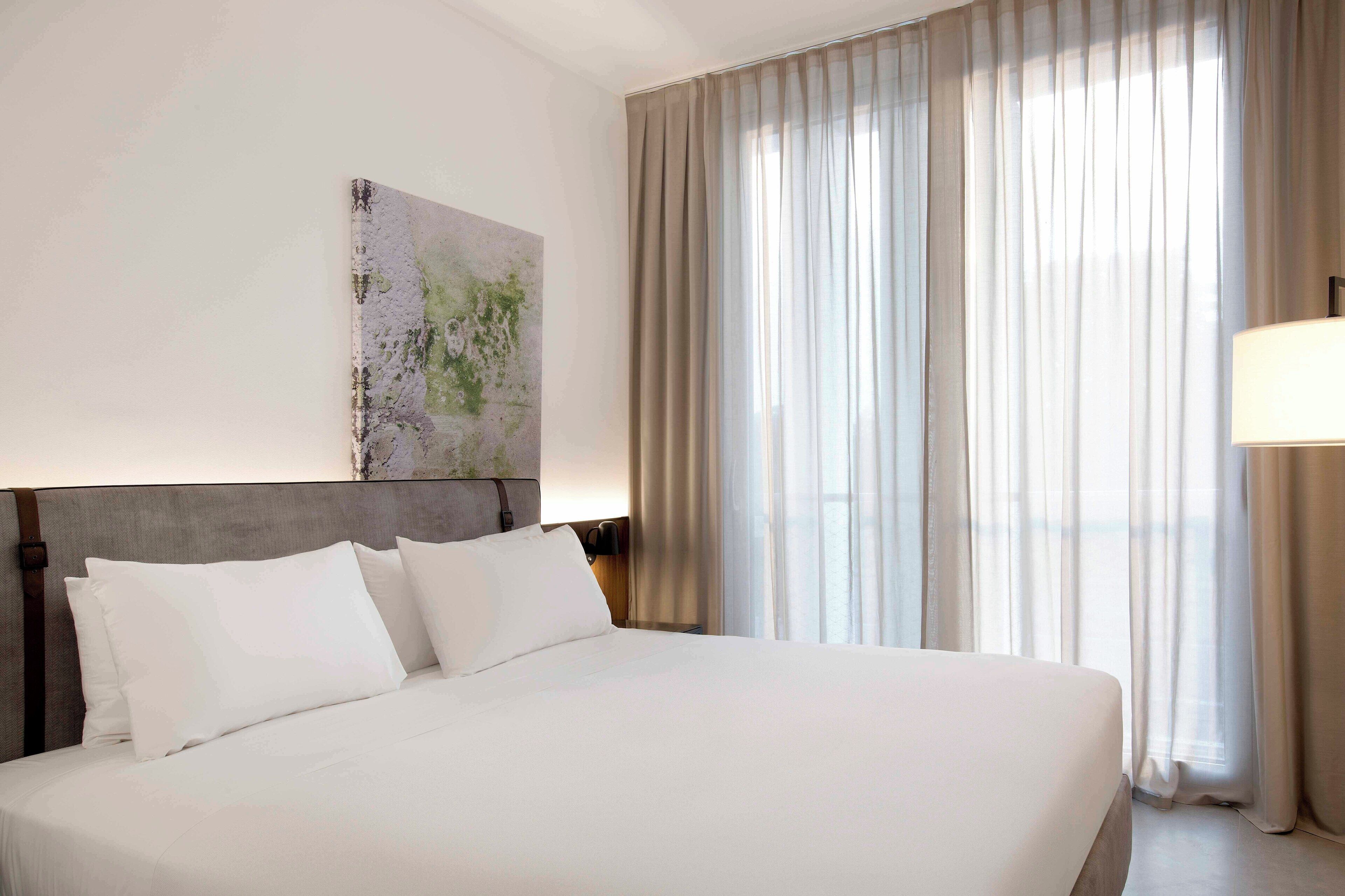 Una habitación de hotel con una cama grande, un cabecero, una obra de arte colgada en la pared y una ventana con cortinas.